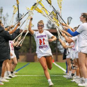 Carly Sullivan runs under a tunnel of lacrosse sticks held aloft by her teammates on her left 和 right. Sullivan has blond hair pulled back 和 wears the white lacrosse uniform with Pomona Pitzer 14 across the top.