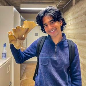 Tommy Shenoi holds up a large native oyster mushroom while st和ing in a lab. Shenoi has medium-length black hair 和 wears a backpack 和 a blue jacket.